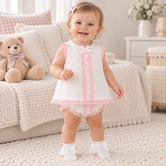 A smiling toddler stands on a cream rug in a soft nursery setting, wearing a white sleeveless dress with pink lace trim and a bow at the neckline, along with white booties. A teddy bear and knitted sofa are visible in the background.