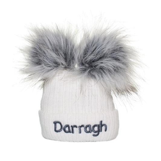 White beanie with gray faux fur pom-poms and 'Darragh' embroidery on a white background