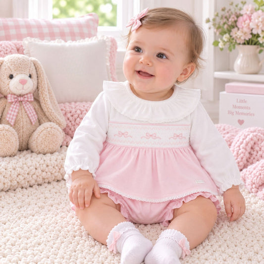 A baby girl sitting on a cream knit blanket in a nursery, wearing a pink and white smocked romper dress with a white ruffled collar and pink ruffled hem. She has a pink hair bow and is surrounded by a plush bunny, pillows, and a knitted blanket.