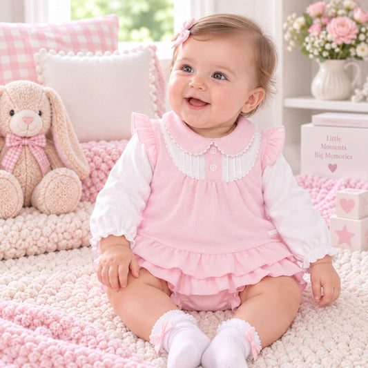 A baby girl sitting on a pink knit blanket in a nursery, wearing a pink ruffled romper over a white long-sleeve shirt. The background features a plush bunny toy, a pillow that reads 'Lullaby Lane Baby Shop', and a small box that says 'Little Moments Big Memories'.