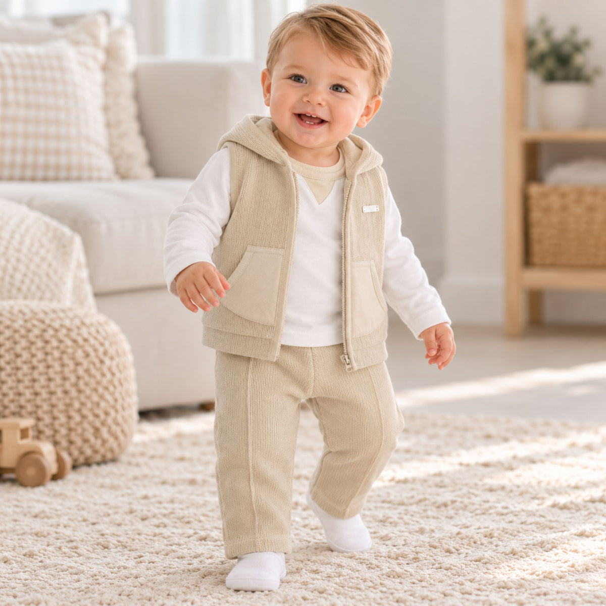 A toddler walking on a shag rug in a bright, modern nursery, wearing a beige corduroy vest and matching pants over a white long-sleeve shirt.