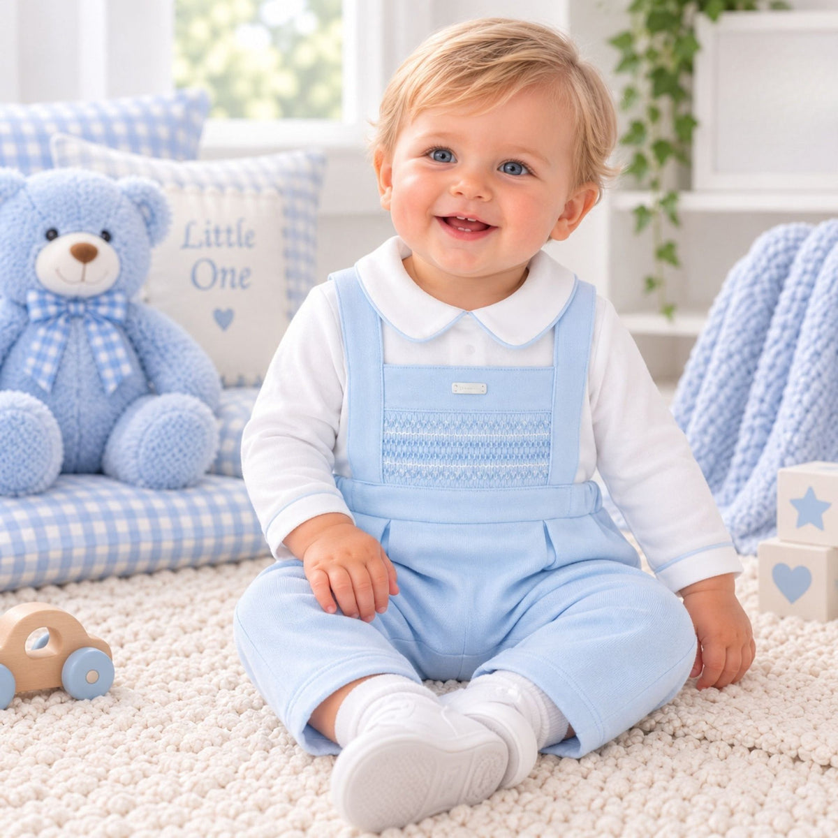 A smiling baby sitting on a cream carpet in a bright nursery, wearing a light blue overall-style romper over a white long-sleeve top. A blue teddy bear and gingham pillows are visible in the background.