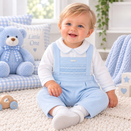 A smiling baby sitting on a cream carpet in a bright nursery, wearing a light blue overall-style romper over a white long-sleeve top. A blue teddy bear and gingham pillows are visible in the background.