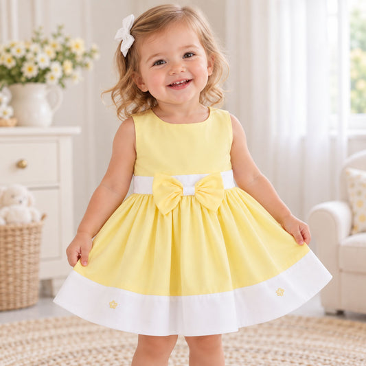 A toddler girl wearing a sleeveless yellow dress with a white waistband, large bow, and white hem trim, standing in a bright nursery-like room.