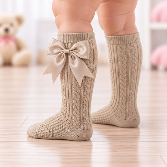 A close-up of a child's legs wearing beige cable-knit socks with a matching satin bow on the front, standing on a light wooden floor with a blurred teddy bear in the background.