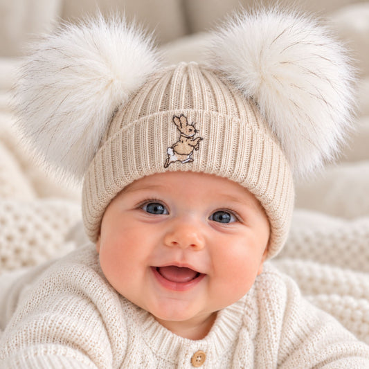 A close-up of a smiling baby wearing a cream ribbed knit beanie with two large white faux-fur pom-poms and a small embroidered bunny on the front, paired with a cream knit sweater.