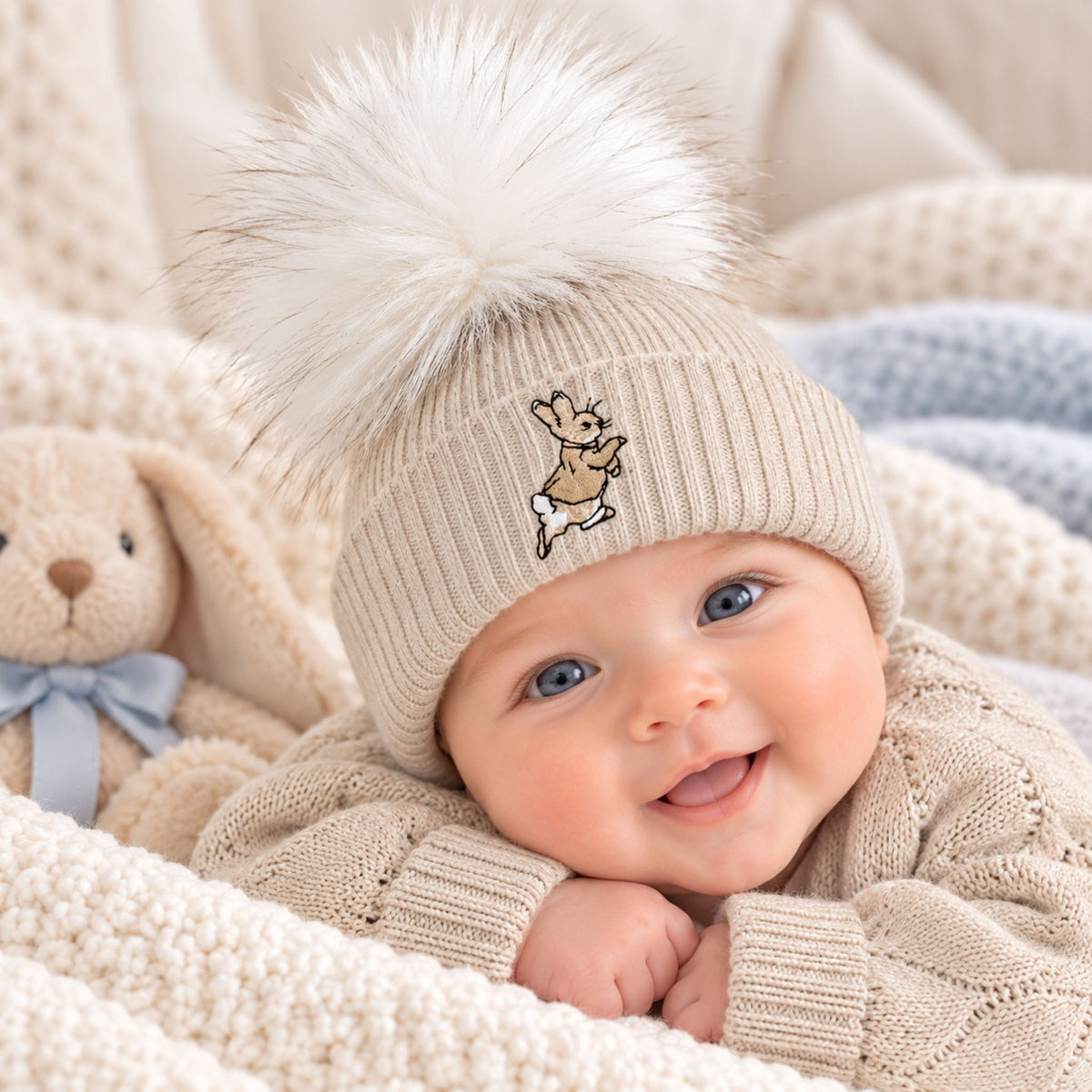 A smiling baby wearing a beige knit beanie with a large white faux-fur pom-pom and a small embroidered bunny motif, lying on cream knit blankets next to a stuffed bunny toy.