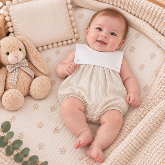 A baby lying on a knitted cream blanket next to a plush stuffed bunny, wearing a cream and beige striped romper with a solid white bib collar.
