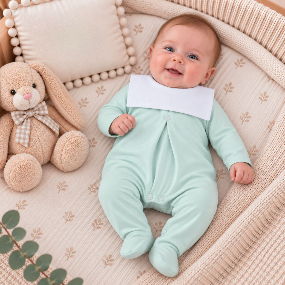A baby lying on a woven bassinet wearing a pale mint green footed sleeper with a contrasting white bib collar. A plush beige bunny toy and a cream pillow are beside the baby.