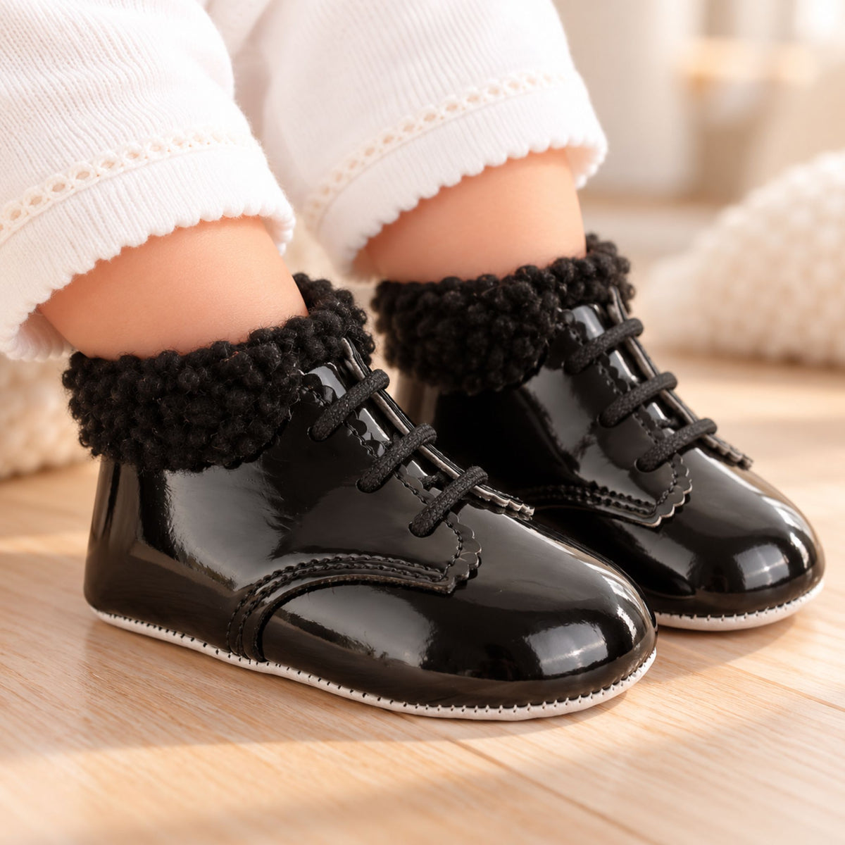A close-up of a baby's feet wearing shiny black lace-up bootis with fluffy black trim, shown on a light wood floor.