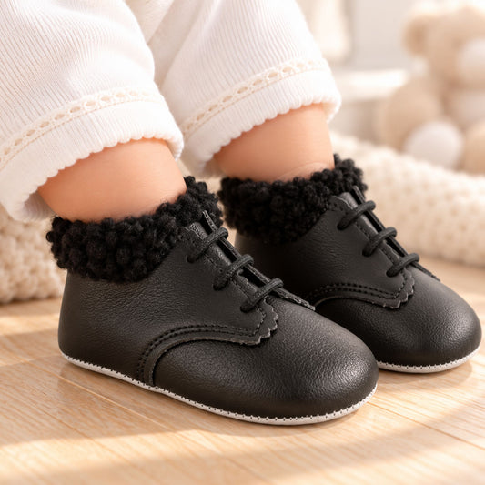 A close-up of a baby's feet wearing black faux-leather lace-up baby shoes with fluffy black knit cuffs, styled with white ribbed socks on a wooden floor.