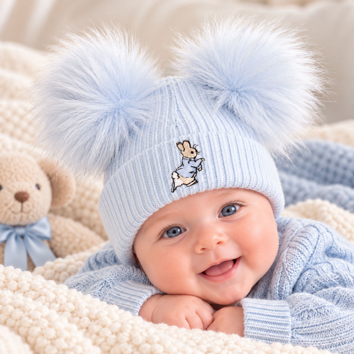 A baby wearing a light blue ribbed knit beanie with two large fluffy pom-poms and a small embroidered bunny character on the front, lying on a cream knit blanket next to a teddy bear.
