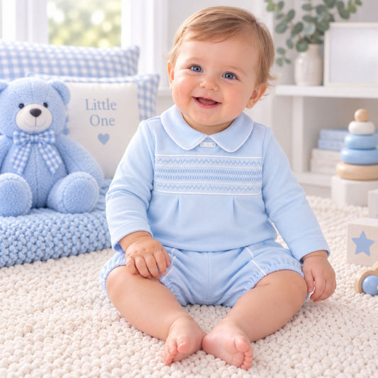 A baby sitting on a cream knit rug wearing a light blue long-sleeve romper with a white Peter Pan collar and decorative horizontal stripes across the chest. The background features a blue teddy bear, a blue and white checkered pillow that says 'Little One', and soft nursery decor.