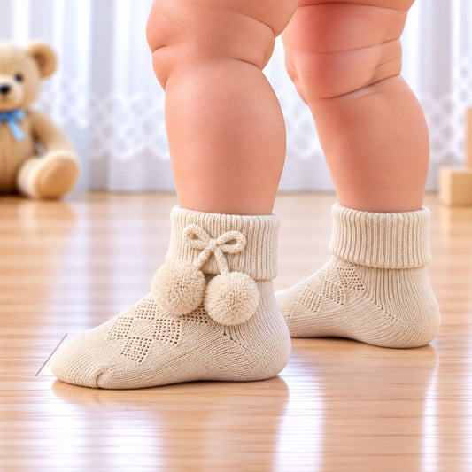 A close-up of a baby's legs wearing beige knit socks with decorative pom-poms and a bow on the front, standing on a wooden floor with a teddy bear in the background.