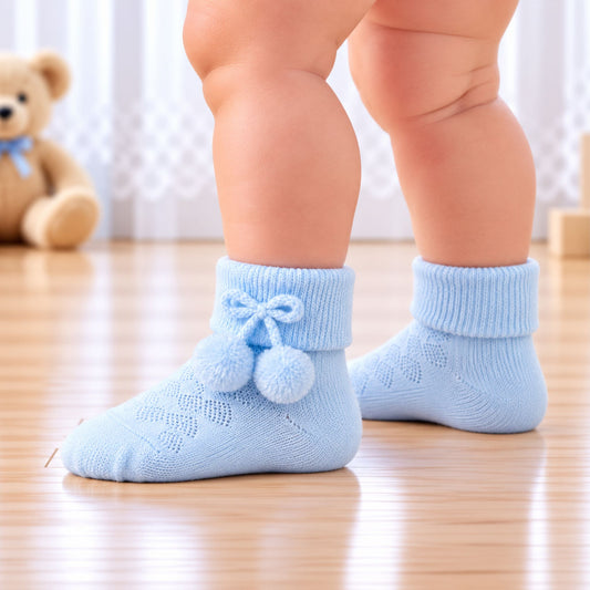 A close-up of a baby's legs wearing light blue knit socks with a bow and pom-pom detail, standing on a wooden floor with a teddy bear in the background.