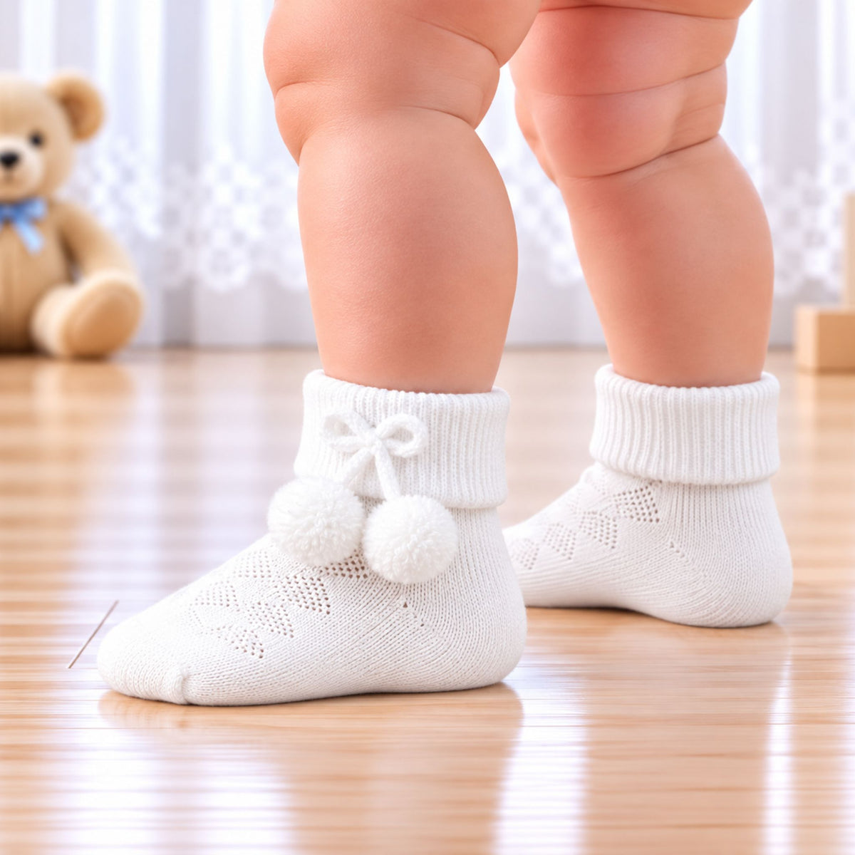 White knit baby socks with pom-pom details worn by a baby standing on a wooden floor, with a teddy bear in the background.