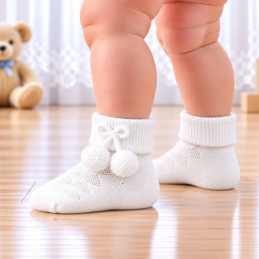 White knit baby socks with pom-pom details worn by a baby standing on a wooden floor, with a teddy bear in the background.