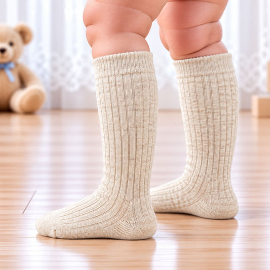 A close-up of a child's legs wearing ribbed beige-colored knee-high socks, standing on a light wooden floor with a soft-focus teddy bear in the background.