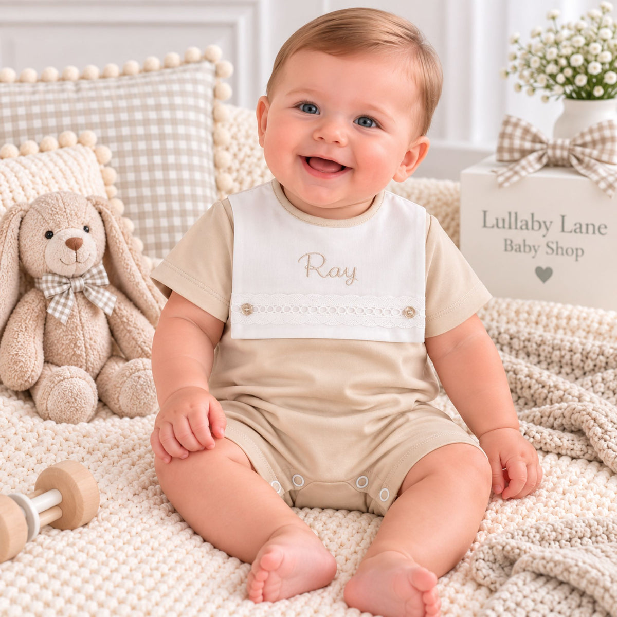 A smiling baby wearing a beige short-sleeve romper with a white bib that says 'Ray' in gold script, sitting on a cream knit blanket in a cozy nursery setting with a stuffed bunny and decorative box in the background.
