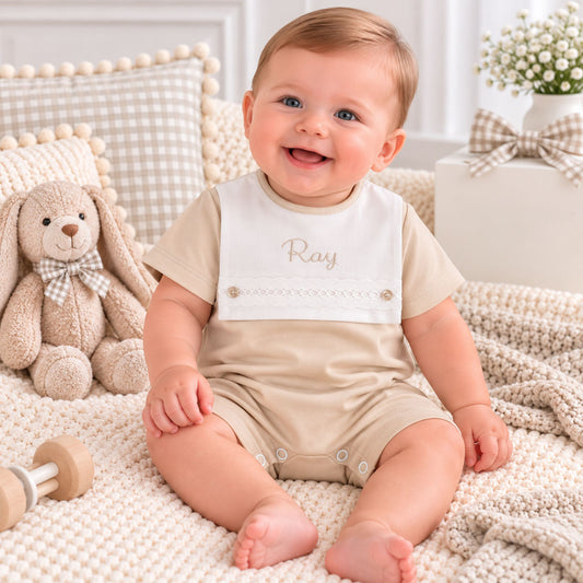 A smiling baby sitting on a cream knit blanket in a styled nursery setting, wearing a beige short-sleeve romper with a white bib panel that has the name 'Ray' embroidered on the chest.