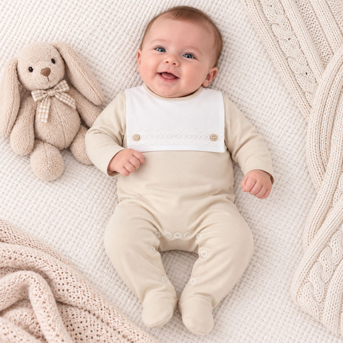 A smiling baby lying on a textured cream blanket next to a plush beige bunny toy, wearing a cream footed sleeper with a white bib panel.