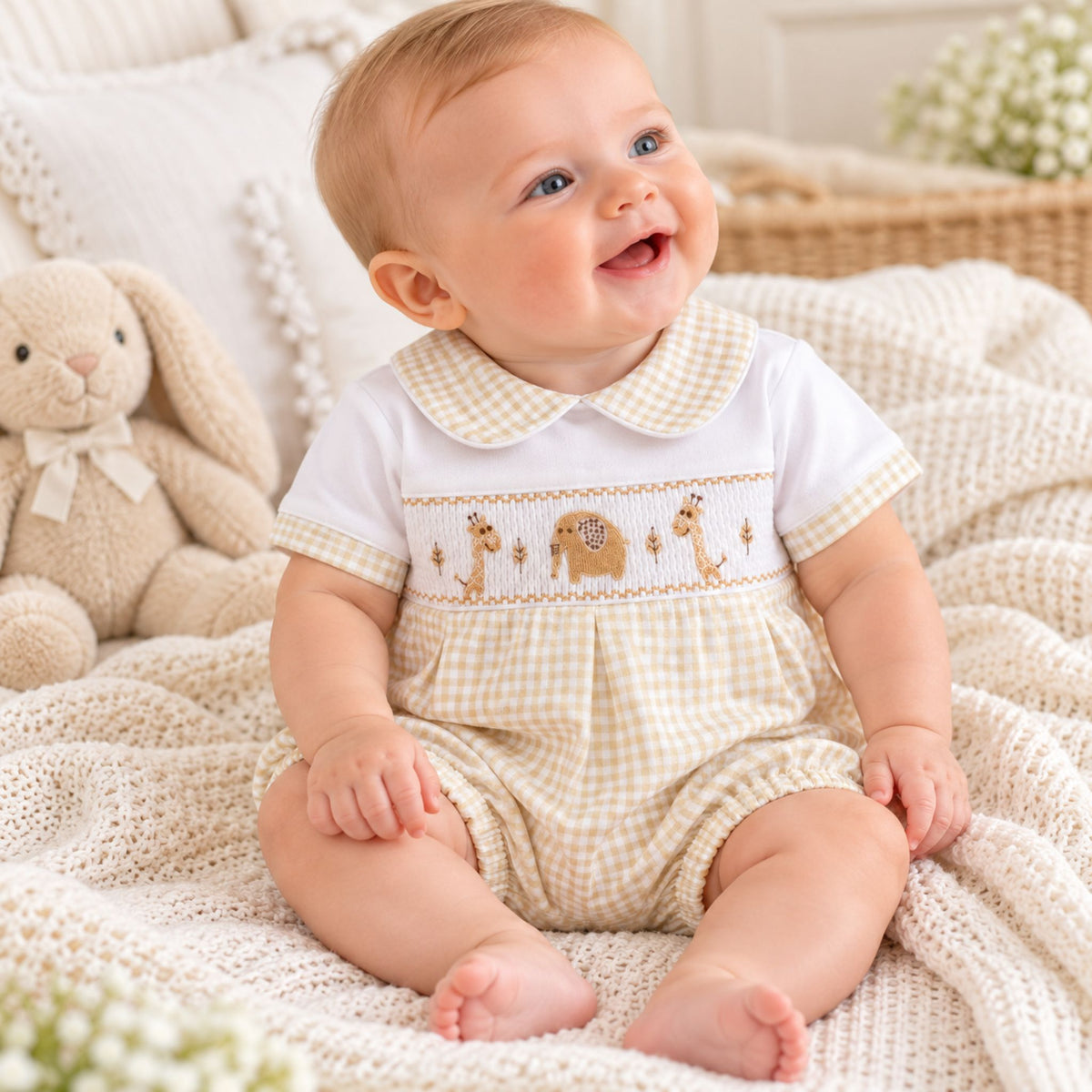A baby sitting on a cream knit blanket wearing a white and beige gingham romper with a smocked giraffe and elephant chest design. A plush bunny toy is visible in the background.
