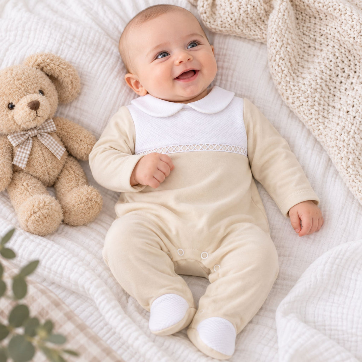 A baby wearing a beige-colored footed sleeper with a white Peter Pan collar and a decorative lace band, lying on a textured white blanket next to a teddy bear.