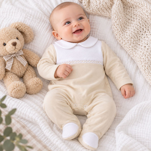 A baby wearing a beige-colored footed sleeper with a white Peter Pan collar and a decorative lace band, lying on a textured white blanket next to a teddy bear.