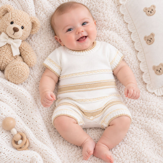 A smiling baby lying on a soft cream knit blanket, wearing a short-sleeve cream knit romper with beige horizontal stripes. A teddy bear toy and a matching pillow are visible around the baby.