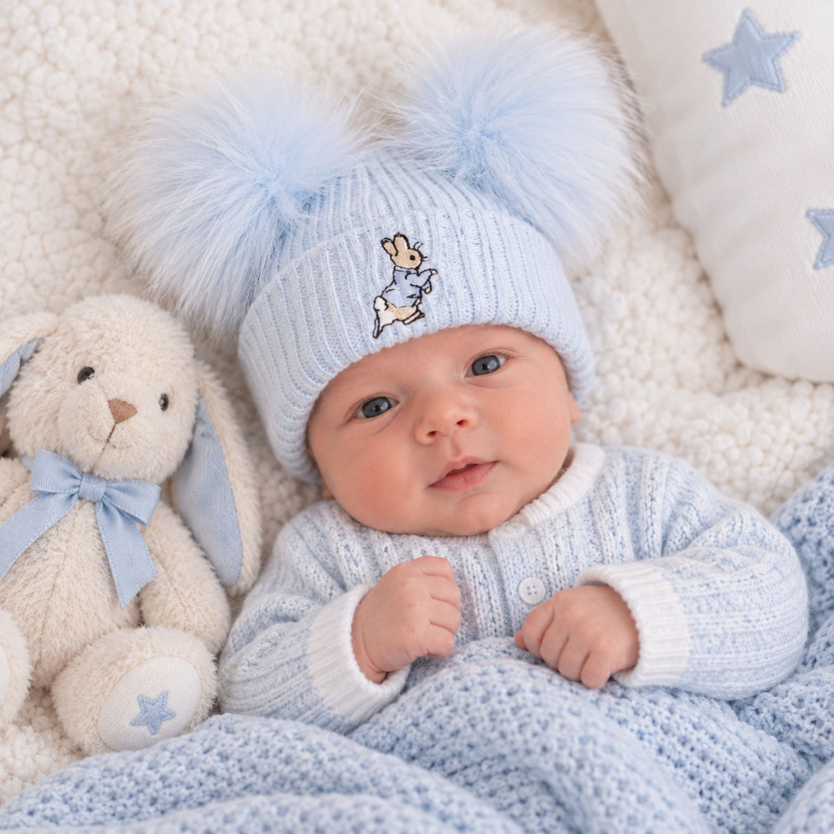 A baby wearing a light blue knit beanie with two large fluffy pom-poms and a small rabbit , lying on a cream textured blanket next to a plush white bunny toy.