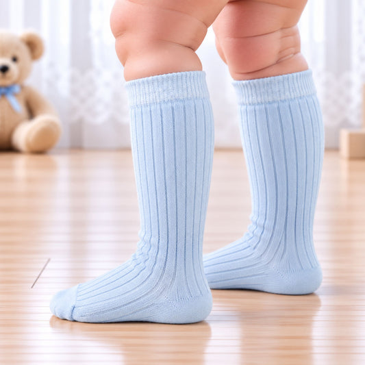 A close-up of a baby's legs wearing light blue ribbed knee-high socks on a wooden floor, with a soft-focus teddy bear in the background.