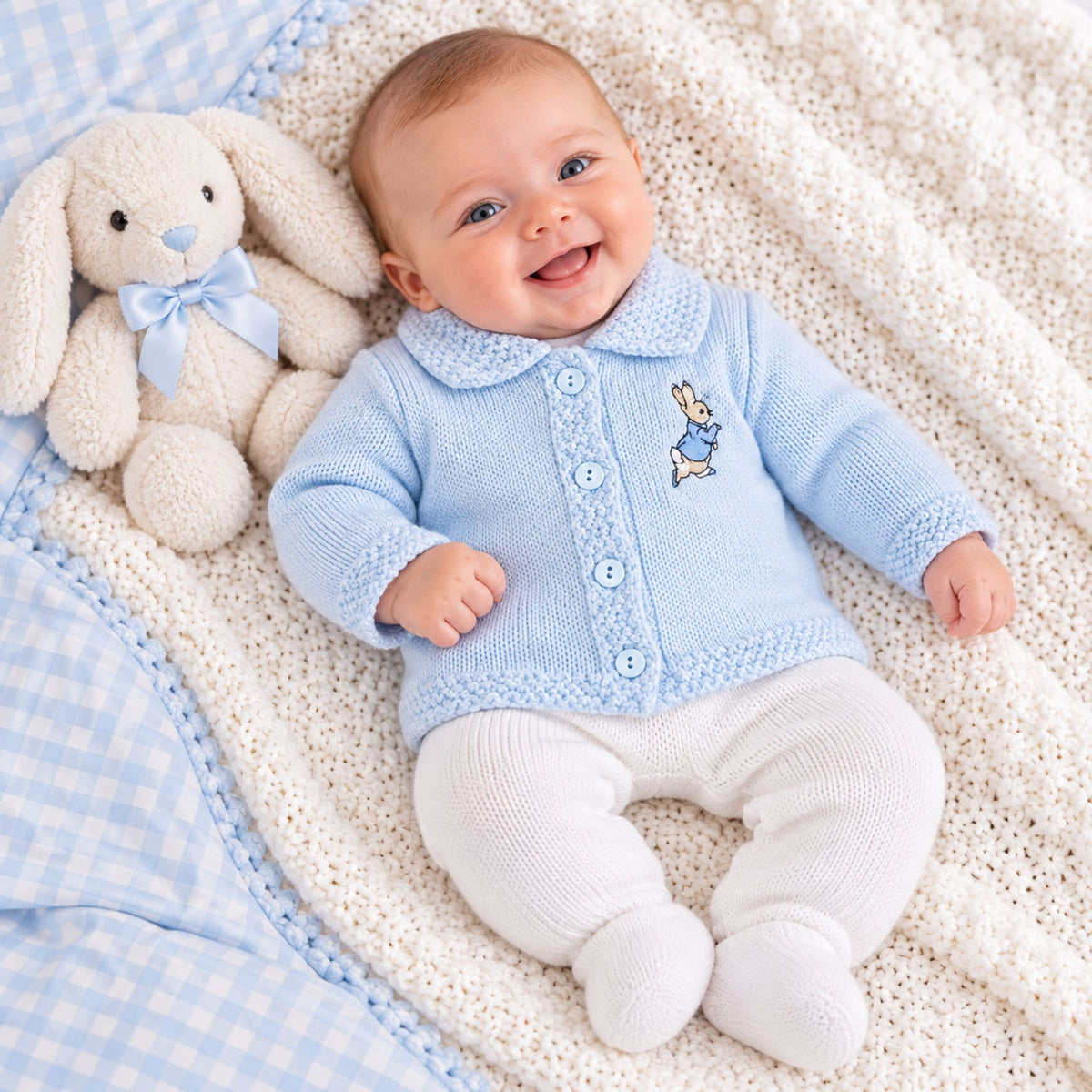A smiling baby lying on a cream knit blanket next to a white bunny toy, wearing a light blue knit cardigan with embroidered rabbit.