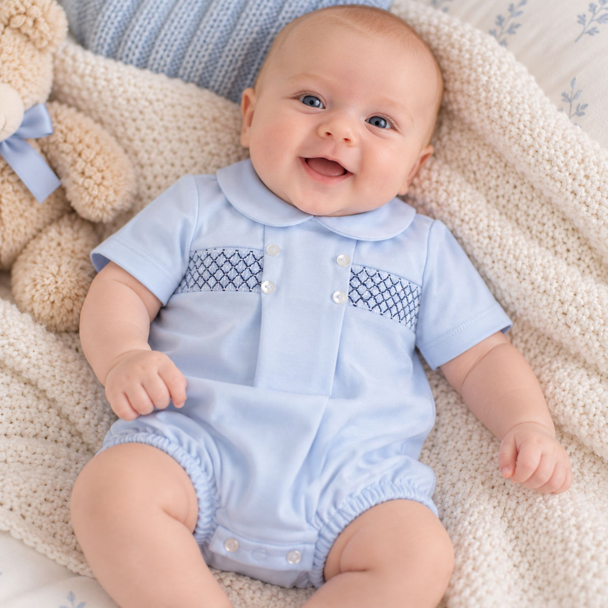 A baby lying on a cream knit blanket next to a teddy bear, wearing a light blue short-sleeve romper with a dark blue patterned chest panel and white snap buttons.