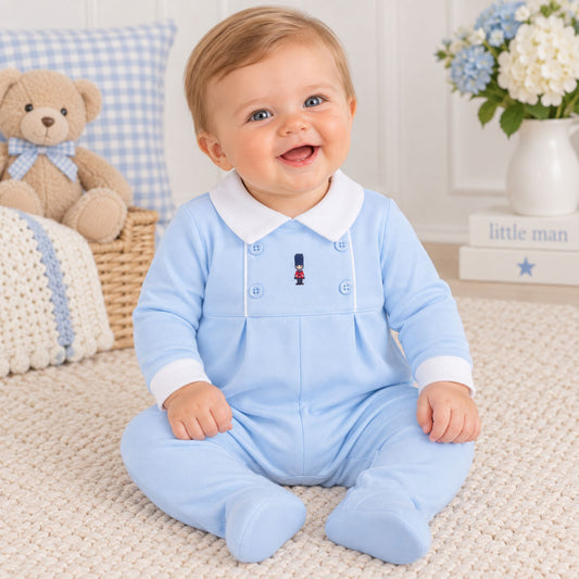 A baby sitting on a cream knit rug wearing a light blue footed romper with a white collar and cuffs. The background features a teddy bear, a blue-and-white check pillow, and a white vase of flowers.