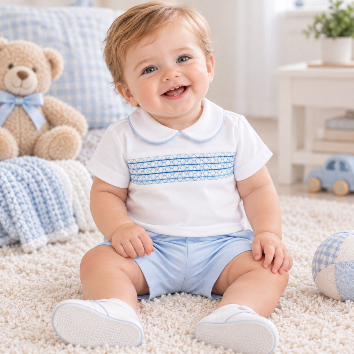 A smiling baby sitting on a cream carpet in a bright nursery, wearing a white short-sleeve Peter Pan collar shirt with a light blue patterned horizontal stripe across the chest and light blue shorts.