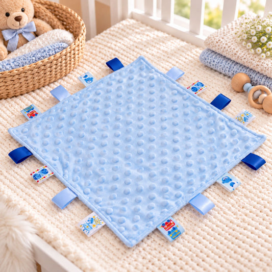 A light blue textured sensory baby blanket with blue fabric tabs, laid out on a cream knit blanket in a nursery setting.