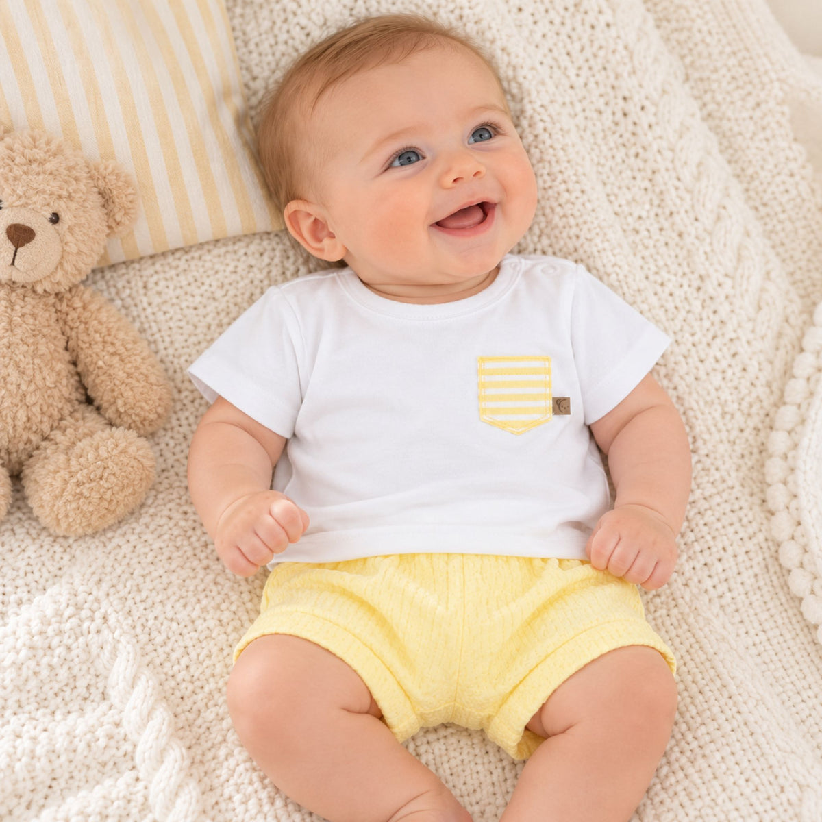 A smiling baby lying on a cream knit blanket next to a teddy bear, wearing a white short-sleeve t-shirt with a yellow striped chest pocket and yellow ribbed shorts.