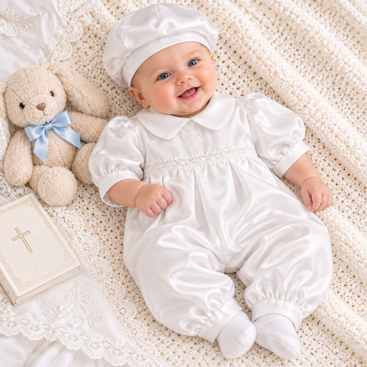 A baby wearing a white satin baptismal outfit with a matching white bonnet lies on a cream crocheted blanket next to a beige teddy bear with a blue bow and a small book with a cross.