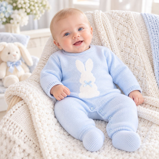 A baby sitting on a cream knit chair wearing a light blue footed romper with a white bunny applique on the chest, surrounded by a cozy cream knit blanket.