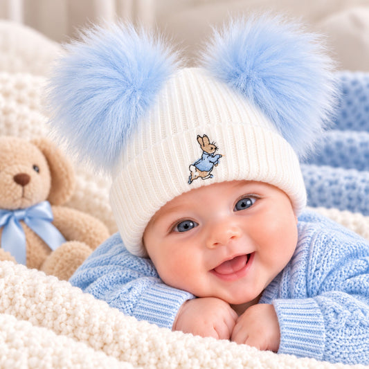 A baby wearing a cream ribbed knit beanie with two fluffy light blue pompoms and a small embroidered bunny  on the front, lying on a soft cream blanket next to a teddy bear.