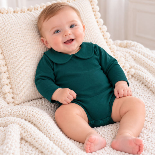 A baby sitting on a cream knit blanket and pillow, wearing a dark green long-sleeve romper with a Peter Pan collar.