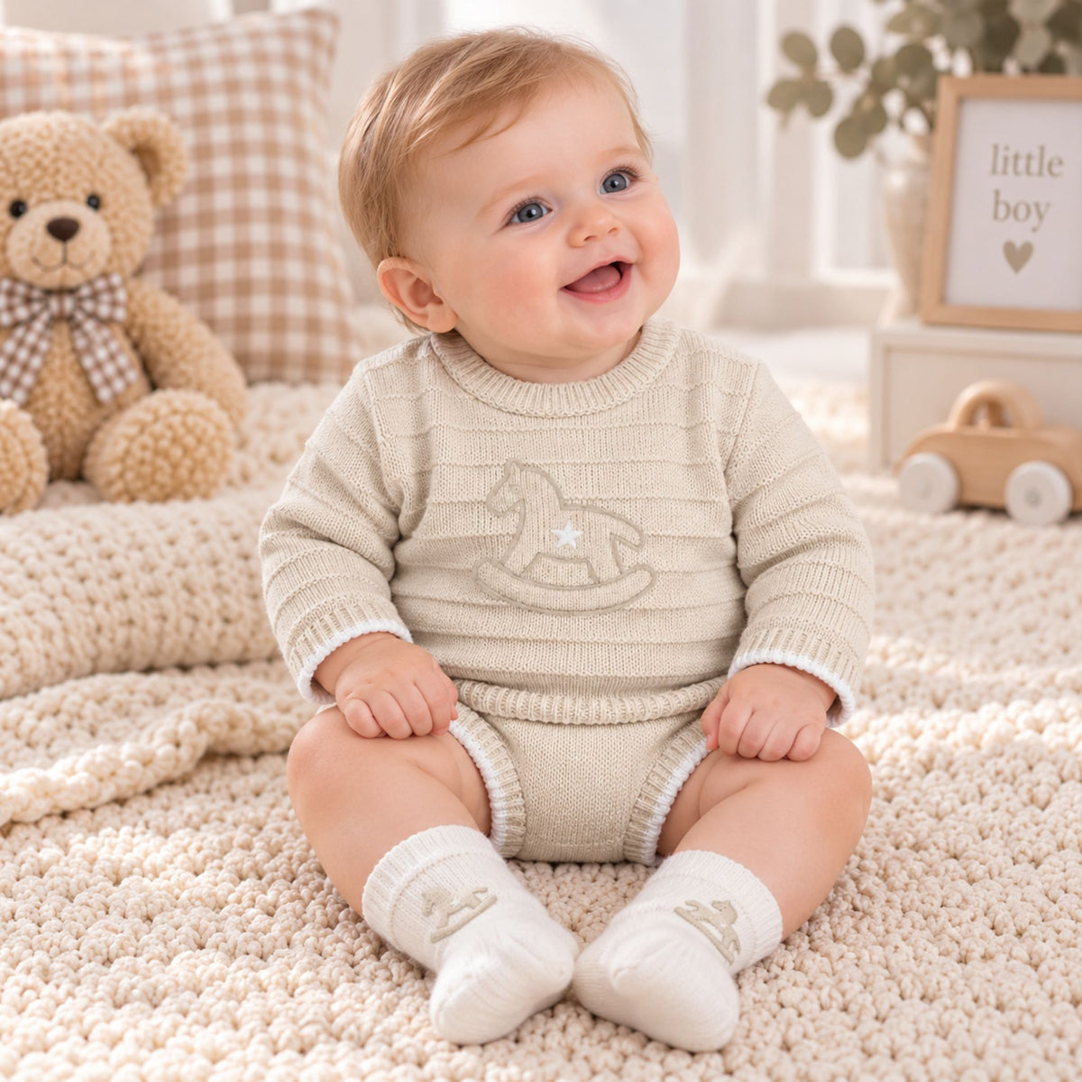A baby sitting on a textured cream rug wearing a ribbed cream knit romper and white socks, with a teddy bear and soft nursery decor in the background.