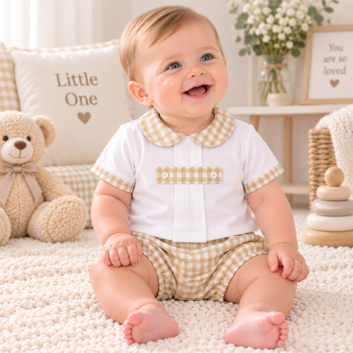 A smiling baby sitting on a cream rug wearing a white short-sleeve top with a beige gingham collar and sleeve trim, paired with matching beige gingham shorts. The background features a teddy bear, a pillow that says 'Little One', and soft nursery decor.