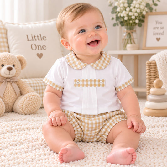 A smiling baby sitting on a cream rug wearing a white short-sleeve top with a beige gingham collar and sleeve trim, paired with matching beige gingham shorts. The background features a teddy bear, a pillow that says 'Little One', and soft nursery decor.