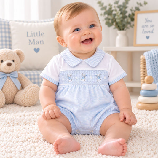 A baby sitting on a white carpet wearing a light blue and white smocked romper, surrounded by baby toys and a teddy bear.