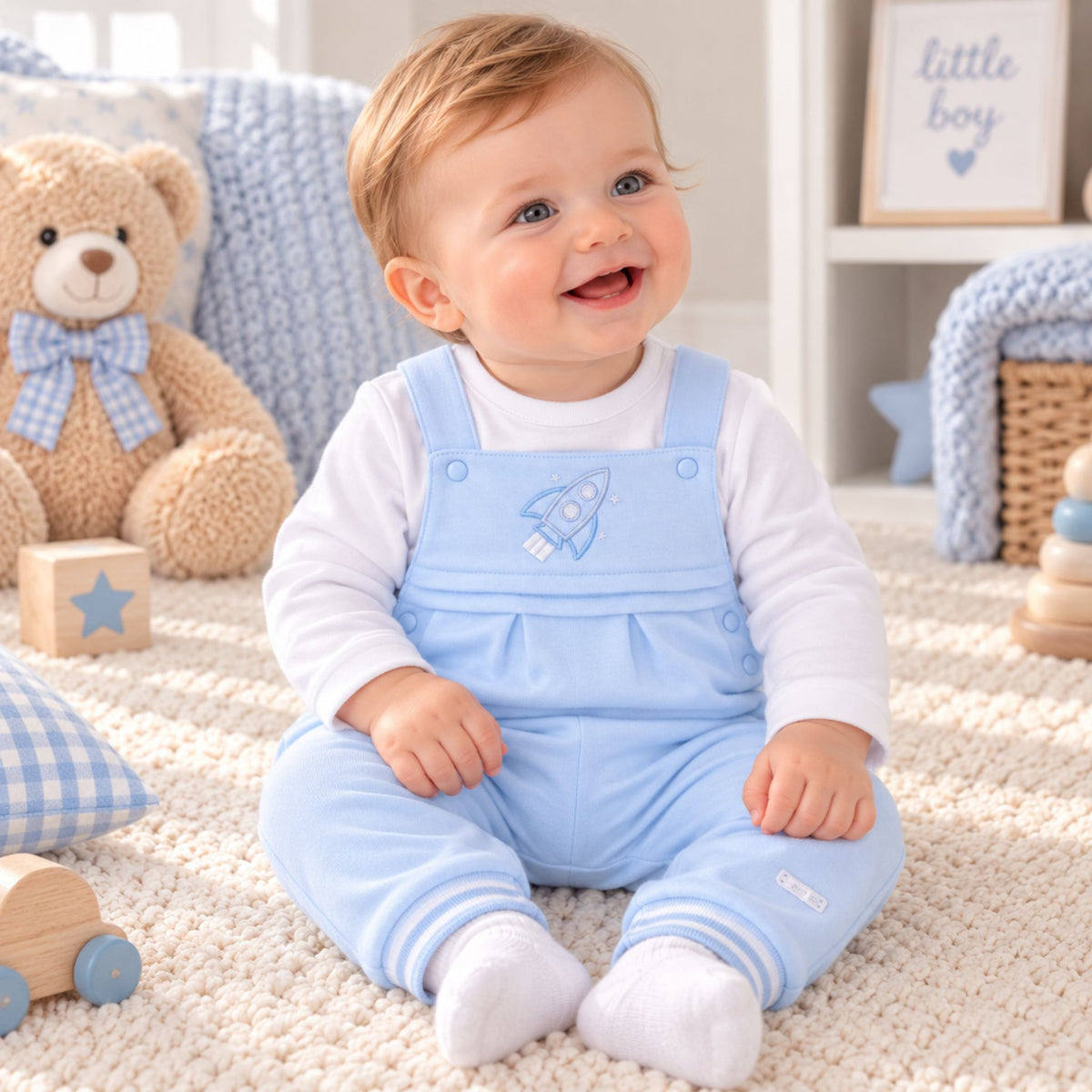 A baby sitting on a cream carpet in a nursery, wearing a light blue overall jumpsuit with a rocket embroidery over a white long-sleeve shirt. A teddy bear, wooden blocks, and soft toys are visible in the background.