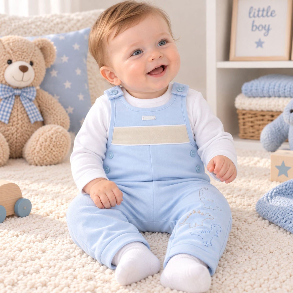 A baby sitting on a cream carpet in a nursery, wearing a light blue footed overalls romper with a white long-sleeve shirt underneath. The background features a plush teddy bear, a blue star-patterned pillow, and soft baby toys.