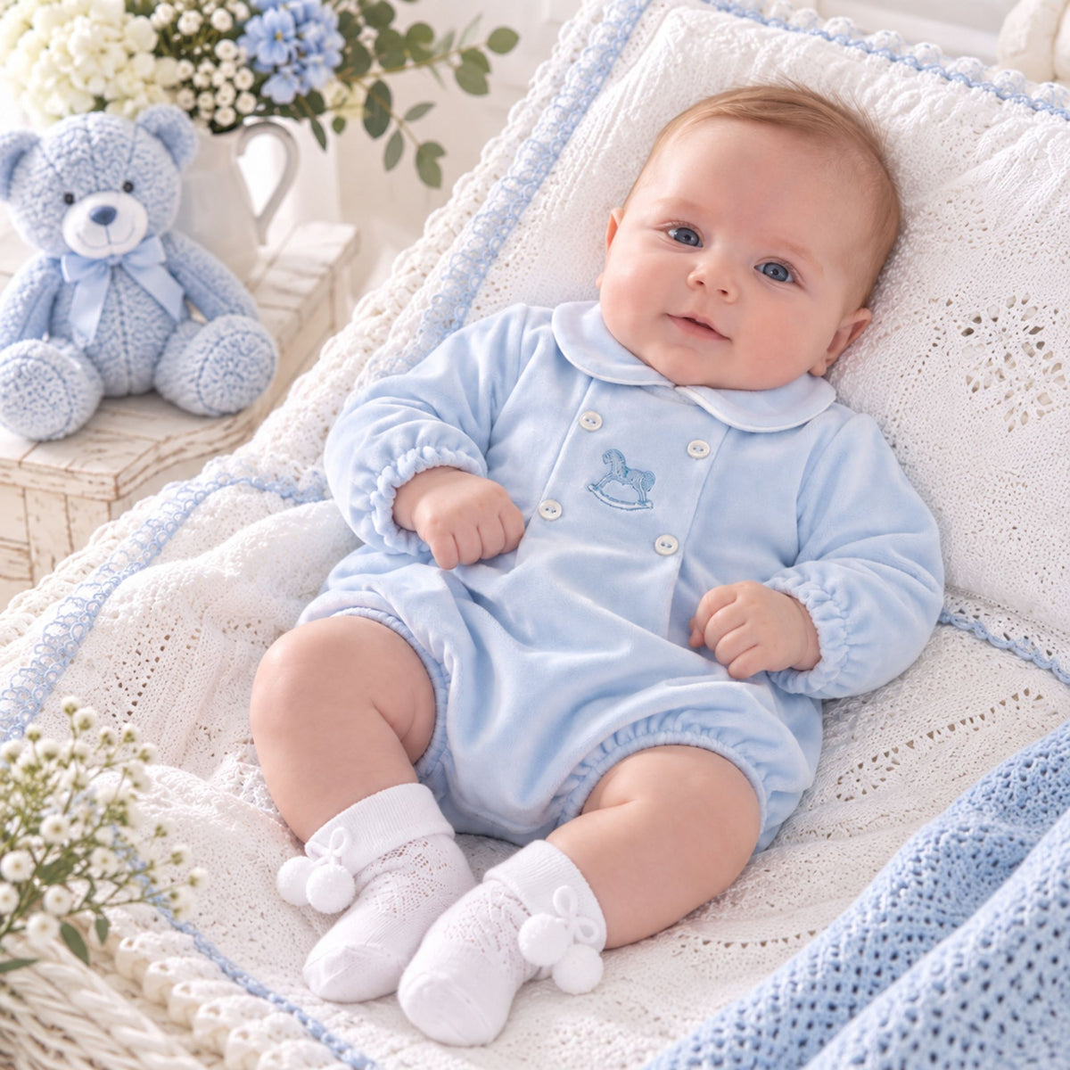 A baby wearing a light blue long-sleeve romper and white socks, lying on a white knitted blanket in a basket with a blue teddy bear and flowers in the background.