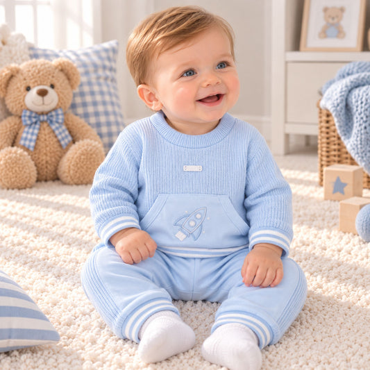 A baby wearing a light blue ribbed knit two-piece outfit with a rocket graphic on the chest, sitting on a cream carpet in a bright nursery.