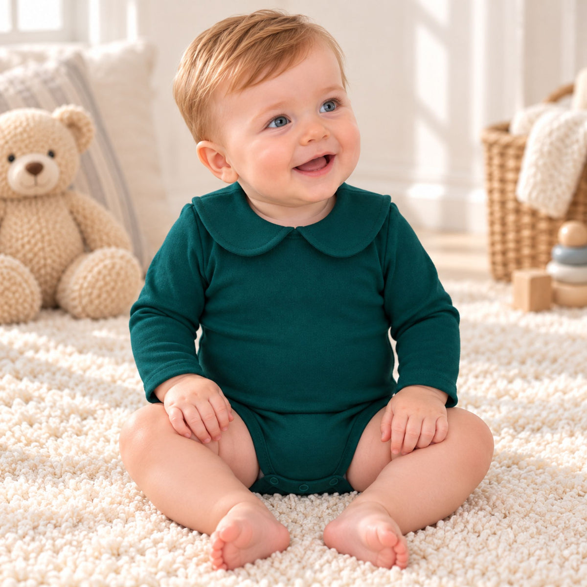 A baby sitting on a white shag rug wearing a dark green long-sleeve romper with a Peter Pan collar, surrounded by a plush teddy bear and soft nursery decor.